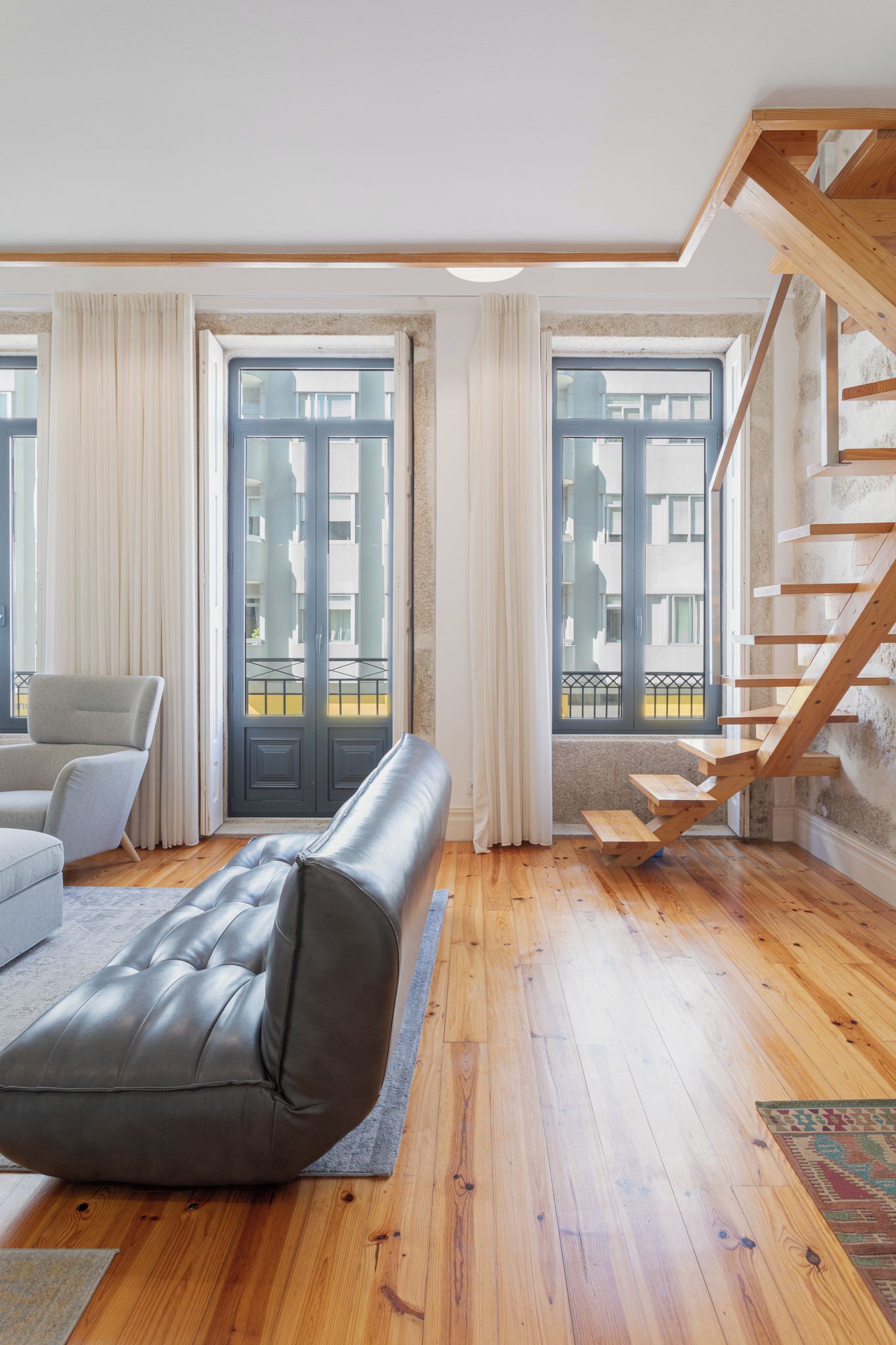 A modern living room with a sofa, a chair, a wooden staircase, and large windows.