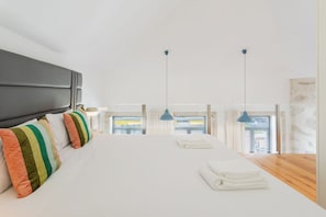 A bedroom with a large bed, a headboard, multiple pillows, two hanging lights, and a view through the window. - Sweet Escape | Charming One-Bedroom Apartment in the Heart of Porto (Porto)