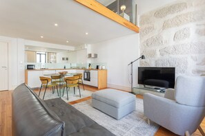 A modern living room with a grey sofa, a grey armchair, and a small ottoman. There is a dining area with a glass table and chairs, and a kitchen with a stone wall. - Sweet Escape | Charming One-Bedroom Apartment in the Heart of Porto (Porto)