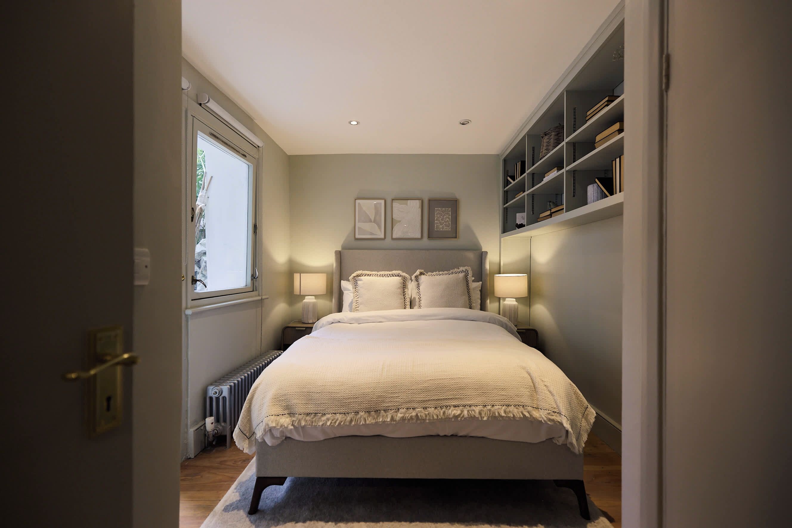 Une chambre à coucher avec un lit, des lampes de chevet, un radiateur, une fenêtre et une bibliothèque.