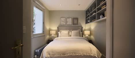 A bedroom with a bed, bedside lamps, a radiator, a window, and a bookshelf.