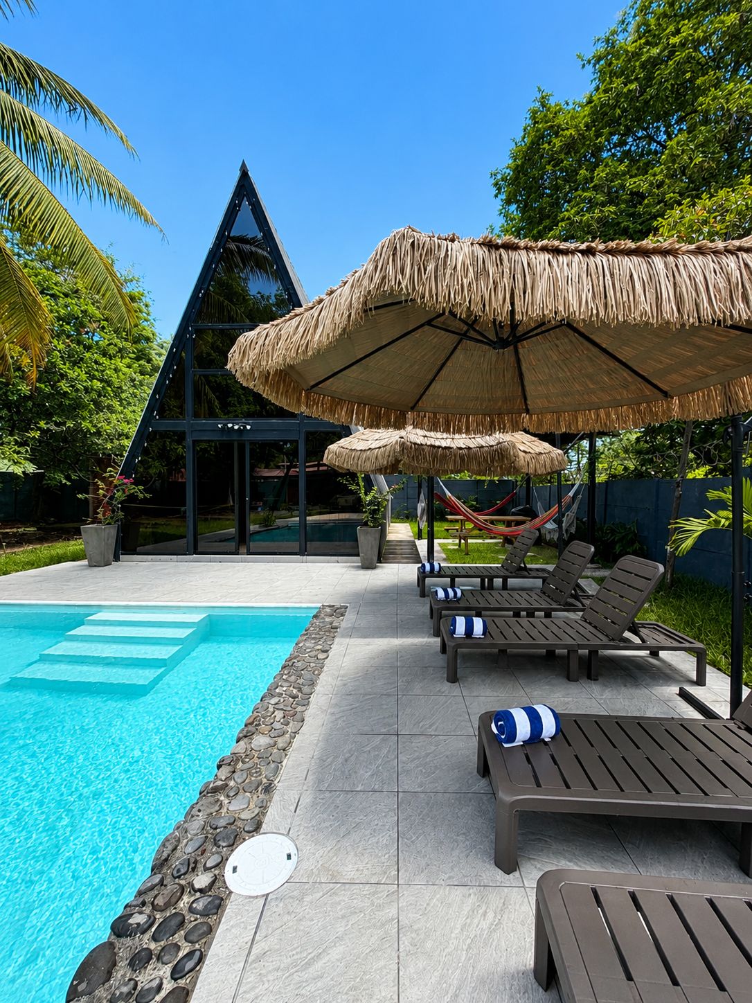 Piscina com espreguiçadeiras e guarda-sóis de palha, cercada por vegetação e uma estrutura de edifício moderna.