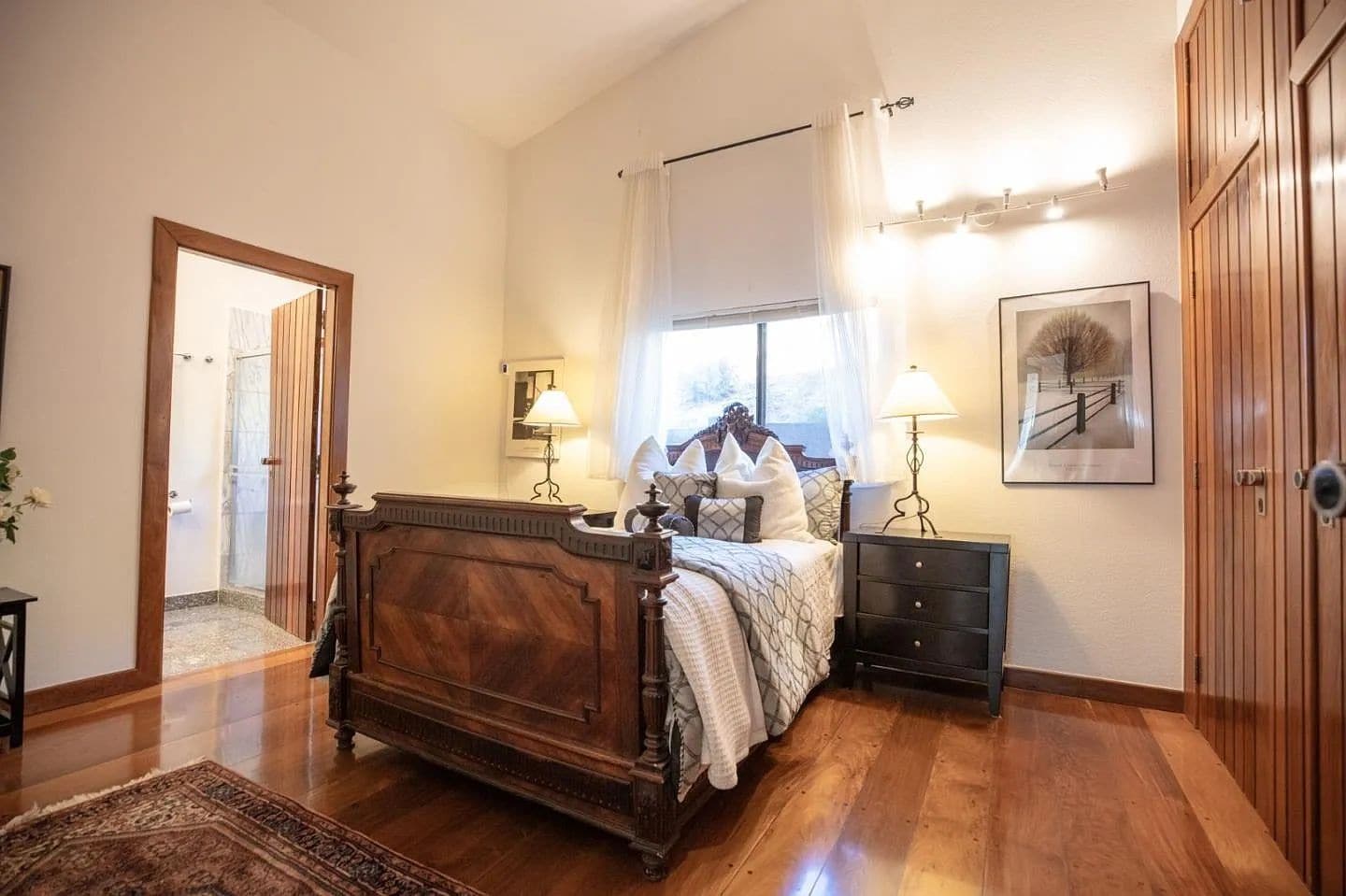 A bedroom with a wooden bed, bedside tables, a nightstand, a window with curtains, a framed picture on the wall, and a door leading to another room.