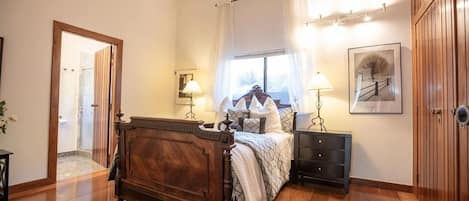 A bedroom with a wooden bed, bedside tables, a nightstand, a window with curtains, a framed picture on the wall, and a door leading to another room.