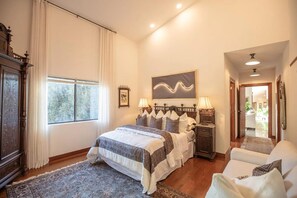A bedroom with a large bed, a nightstand, a chair, a window with curtains, a painting on the wall, and a hallway leading to an outdoor area.