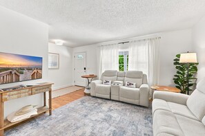 A living room with a sofa, a television, a lamp, and a window with curtains. - Little Minnow - 3 Bedrooms - Mainland (Pawleys Island)