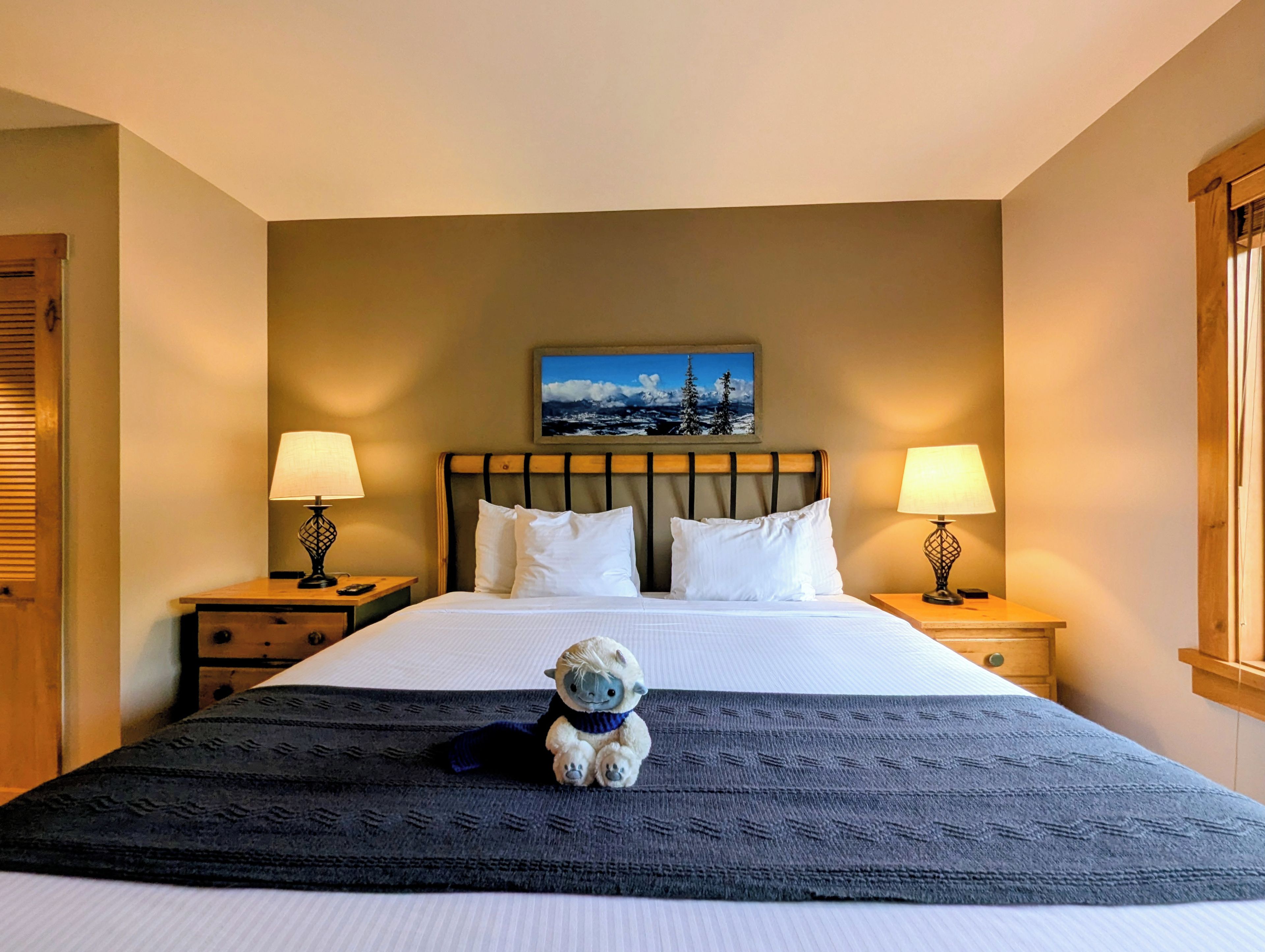 A hotel room with a bed, two bedside tables, a nightstand, a lamp, a painting on the wall, and a stuffed toy on the bed.