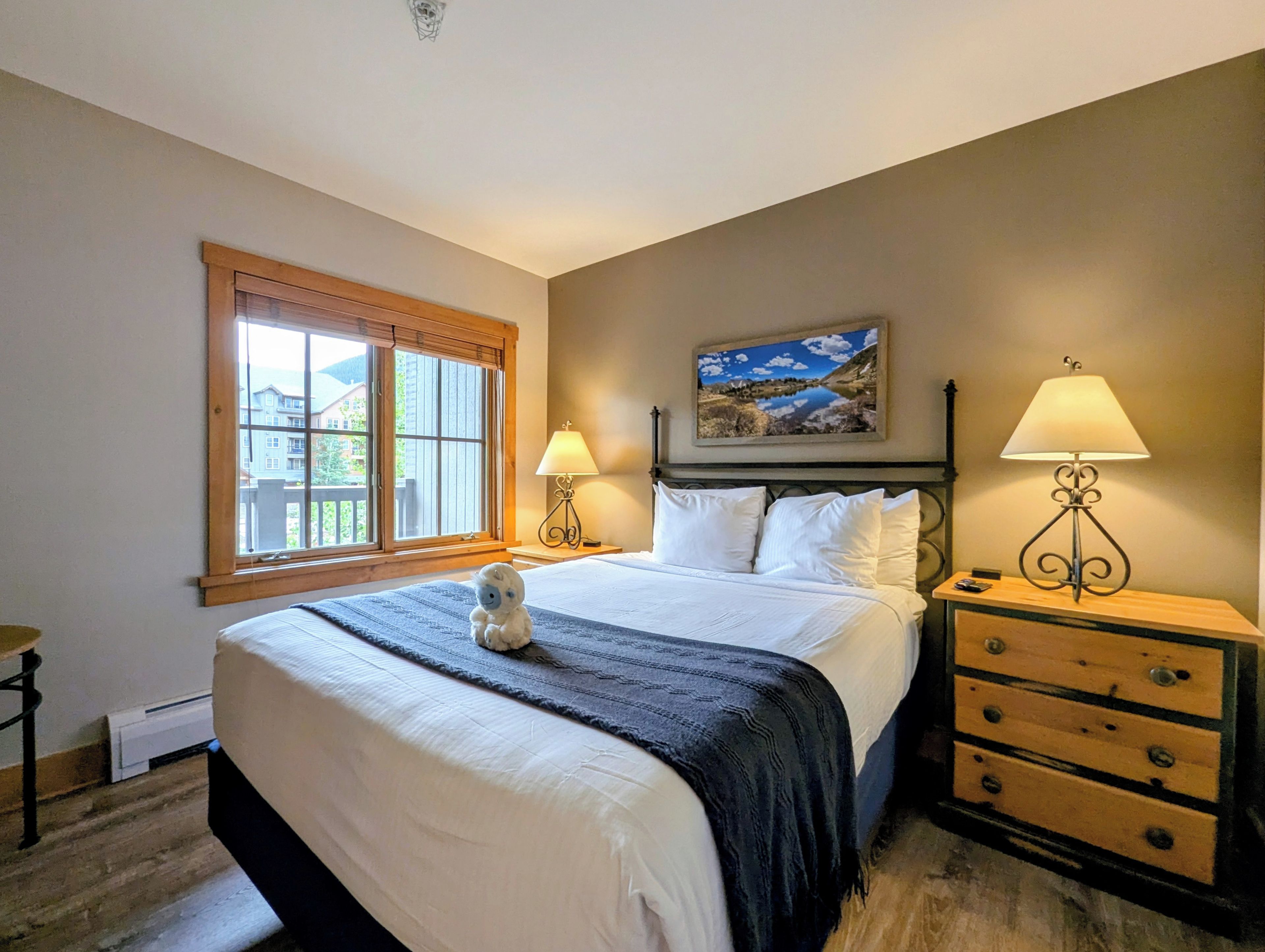 Une chambre à coucher avec un lit, deux lampes de chevet, une table de chevet, une commode, une fenêtre avec des stores et un tableau accroché au mur.