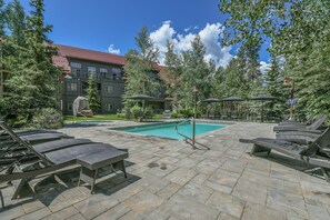 A pool area with lounge chairs and umbrellas, surrounded by trees and a building with a red roof. - Prime Location | Steps to Slopes | King Bed (Keystone)