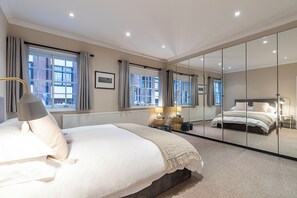 Een moderne slaapkamer met een groot bed, een nachtkastje met lampen, een spiegel en uitzicht naar buiten door grote ramen.