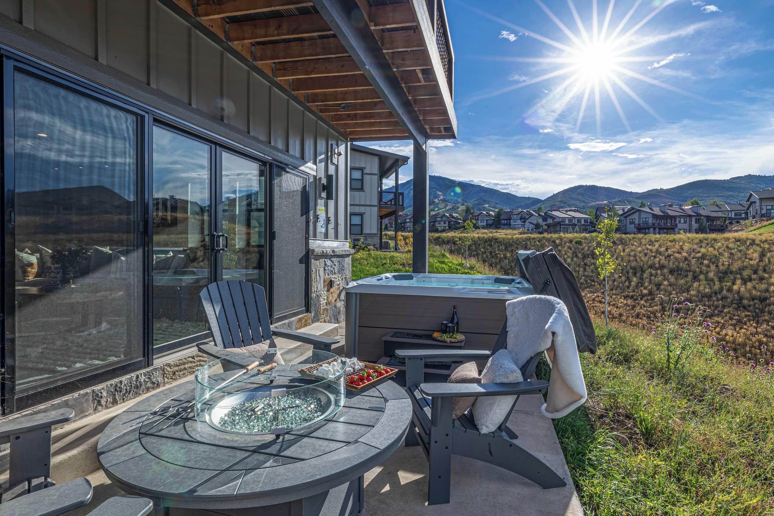 A modern outdoor patio with a glass-top table, chairs, a hot tub, and a scenic mountain view.