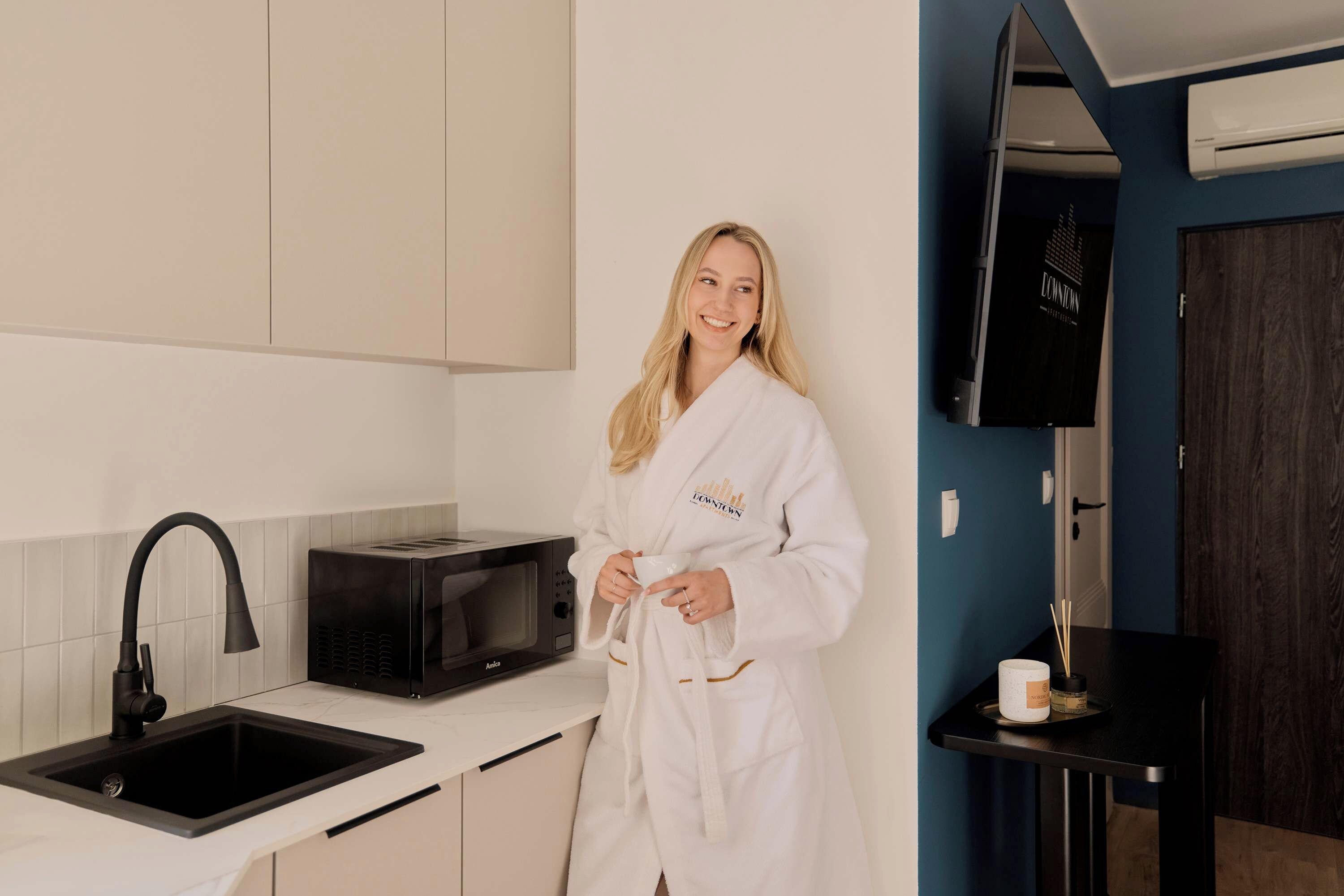 A person in a white robe standing in a modern kitchen with a sink, a black microwave, and a black faucet.