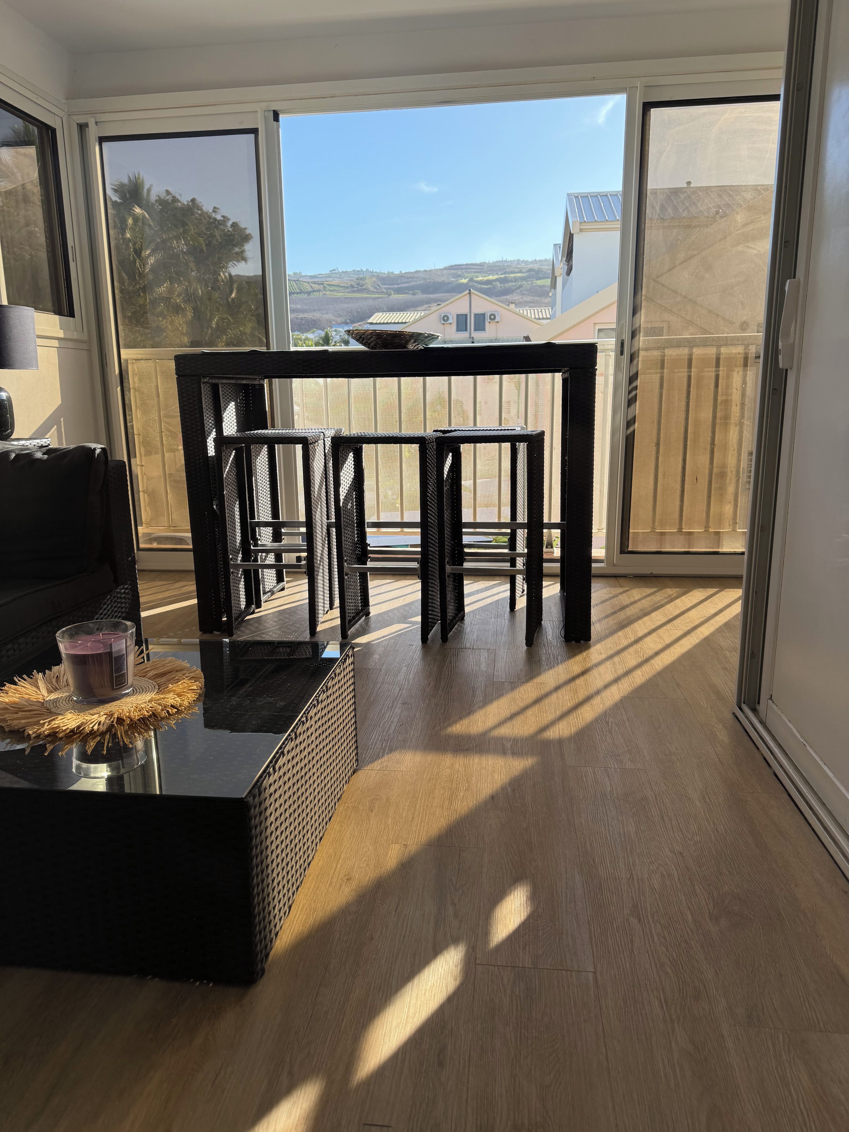 A room with a black wicker table and chairs, a glass coffee table with a candle, and a view through a sliding glass door to the outside.