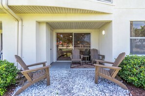 Ein überdachter Patio-Bereich mit Adirondack-Stühlen und einem Tisch, eine Schiebetür, die in einen Garten führt, und ein kleines Fenster mit Jalousien.