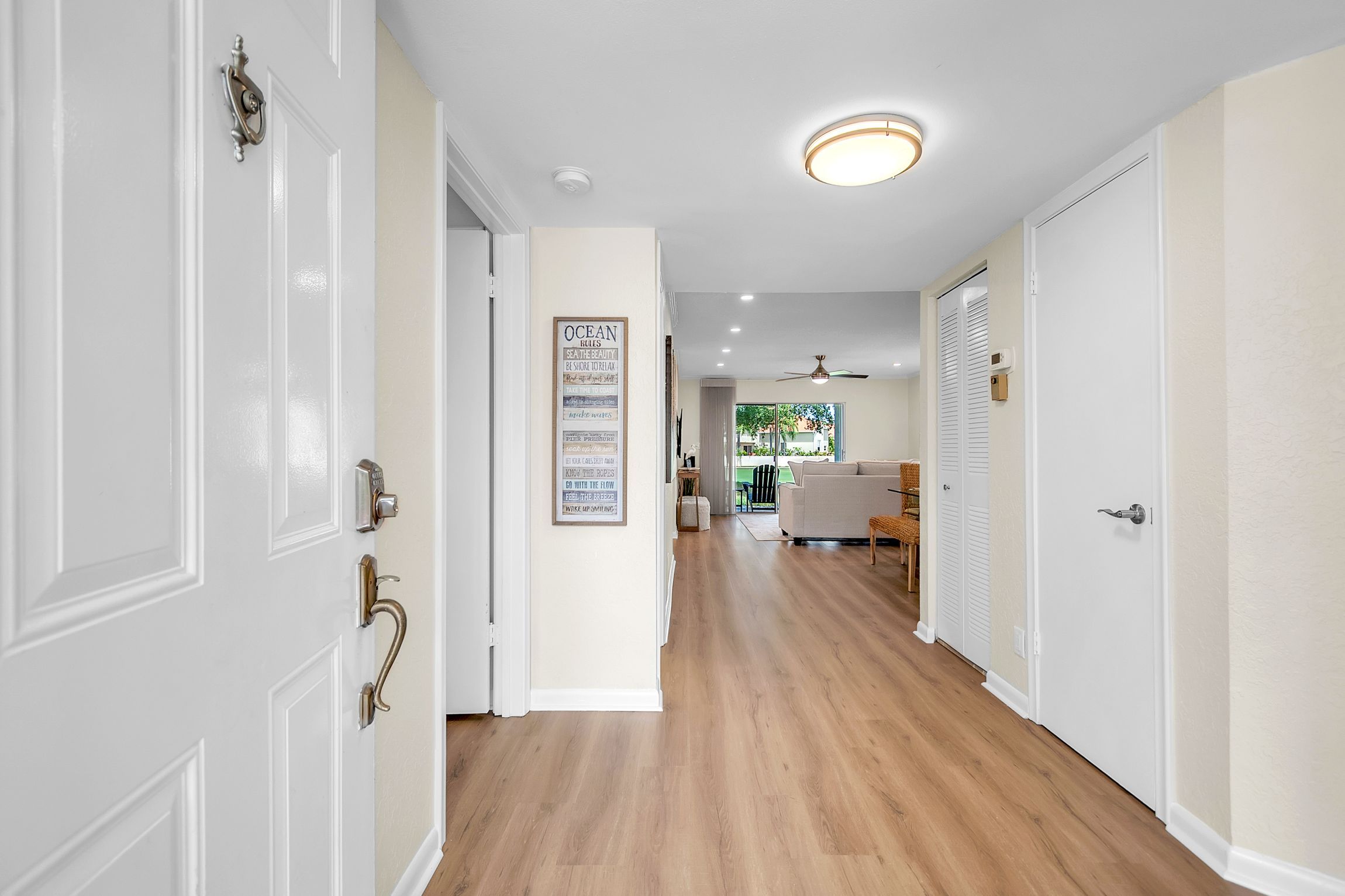 Un couloir avec des portes blanches, un ventilateur de plafond, des spots encastrés et une vue sur un espace de vie comprenant un canapé et une table à manger.