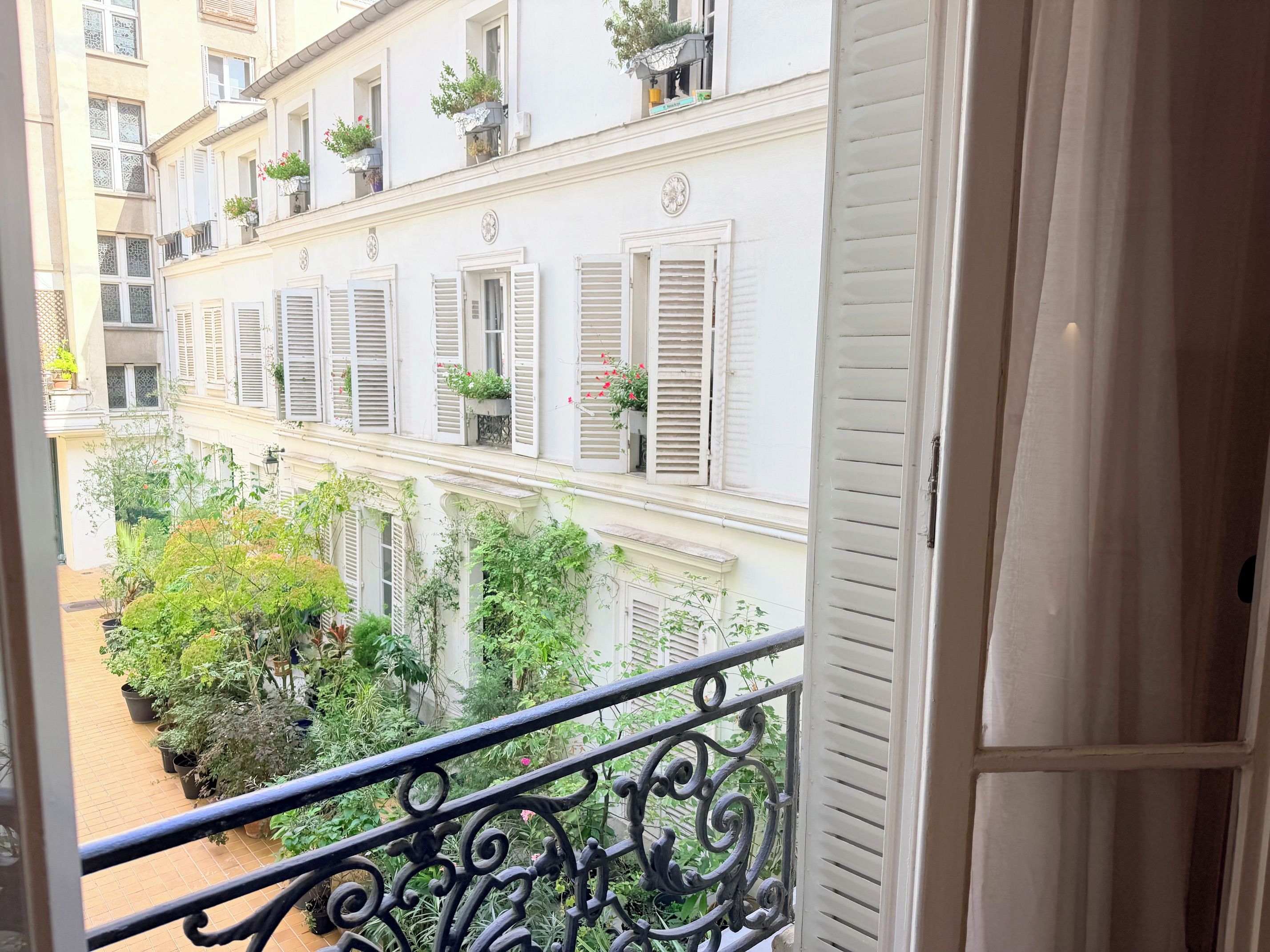 Un balcon orné de balustrades en fer forgé, donnant sur une cour intérieure agrémentée de plantes en pot et sur un bâtiment aux volets blancs.
