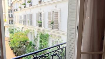 Un balcon orné de balustrades en fer forgé, donnant sur une cour intérieure agrémentée de plantes en pot et sur un bâtiment aux volets blancs.