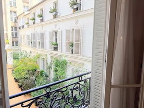 Un balcon orné de balustrades en fer forgé, donnant sur une cour intérieure agrémentée de plantes en pot et sur un bâtiment aux volets blancs.
