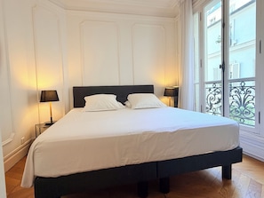 Un lit avec du linge de lit blanc, deux lampes de chevet et une vue sur l’extérieur par une fenêtre donnant sur un balcon en fer forgé.