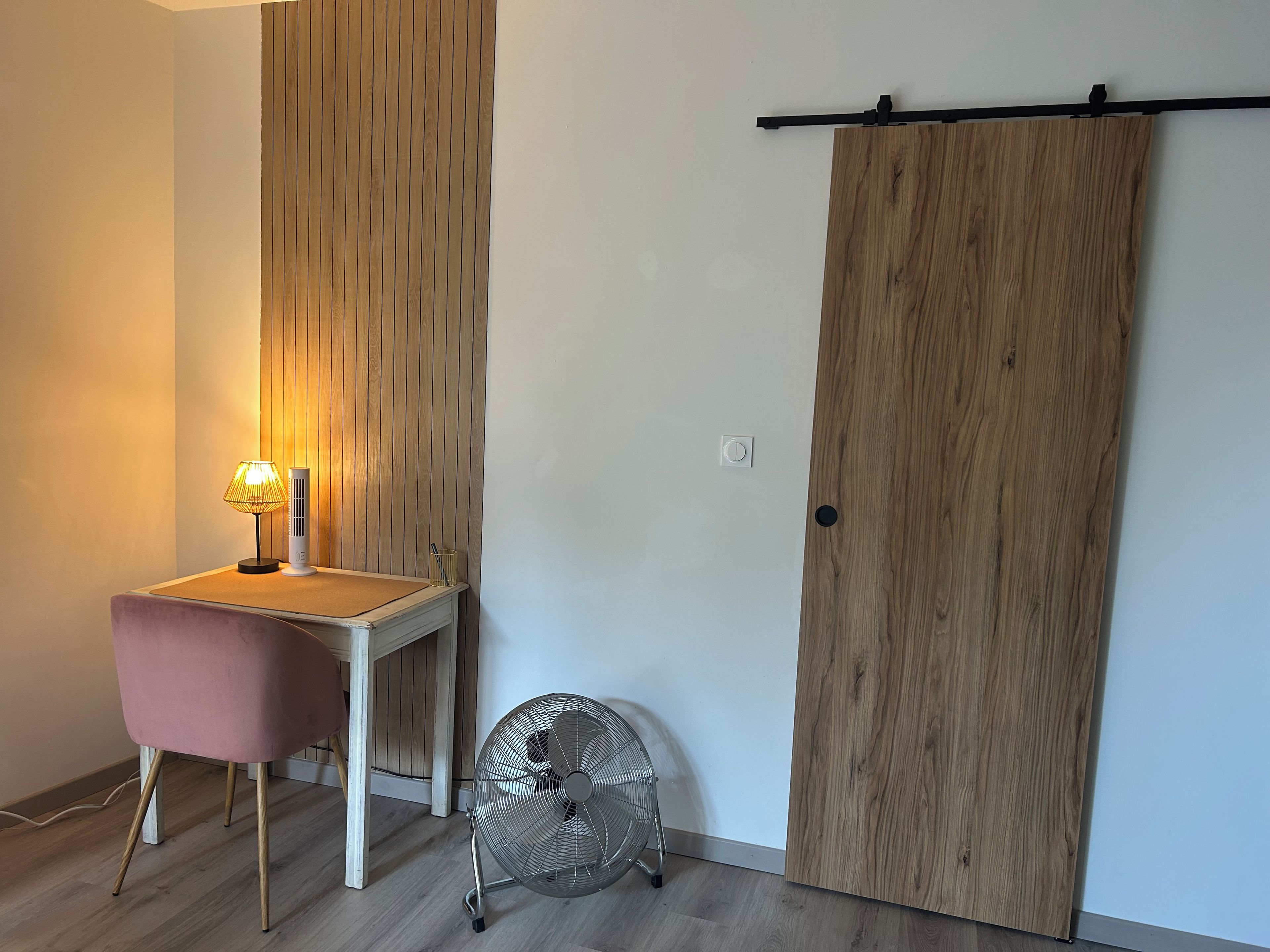 Ein Zimmer mit Schreibtisch, Stuhl, Standventilator und einer Schiebetür.