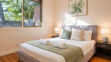 A neatly made bed with a green throw blanket and two pillows, a bedside table with a lamp, and a window with a view of trees.