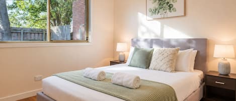 Een netjes opgemaakt bed met een groene deken en twee kussens, een nachtkastje met een lamp, en een raam met uitzicht op bomen.