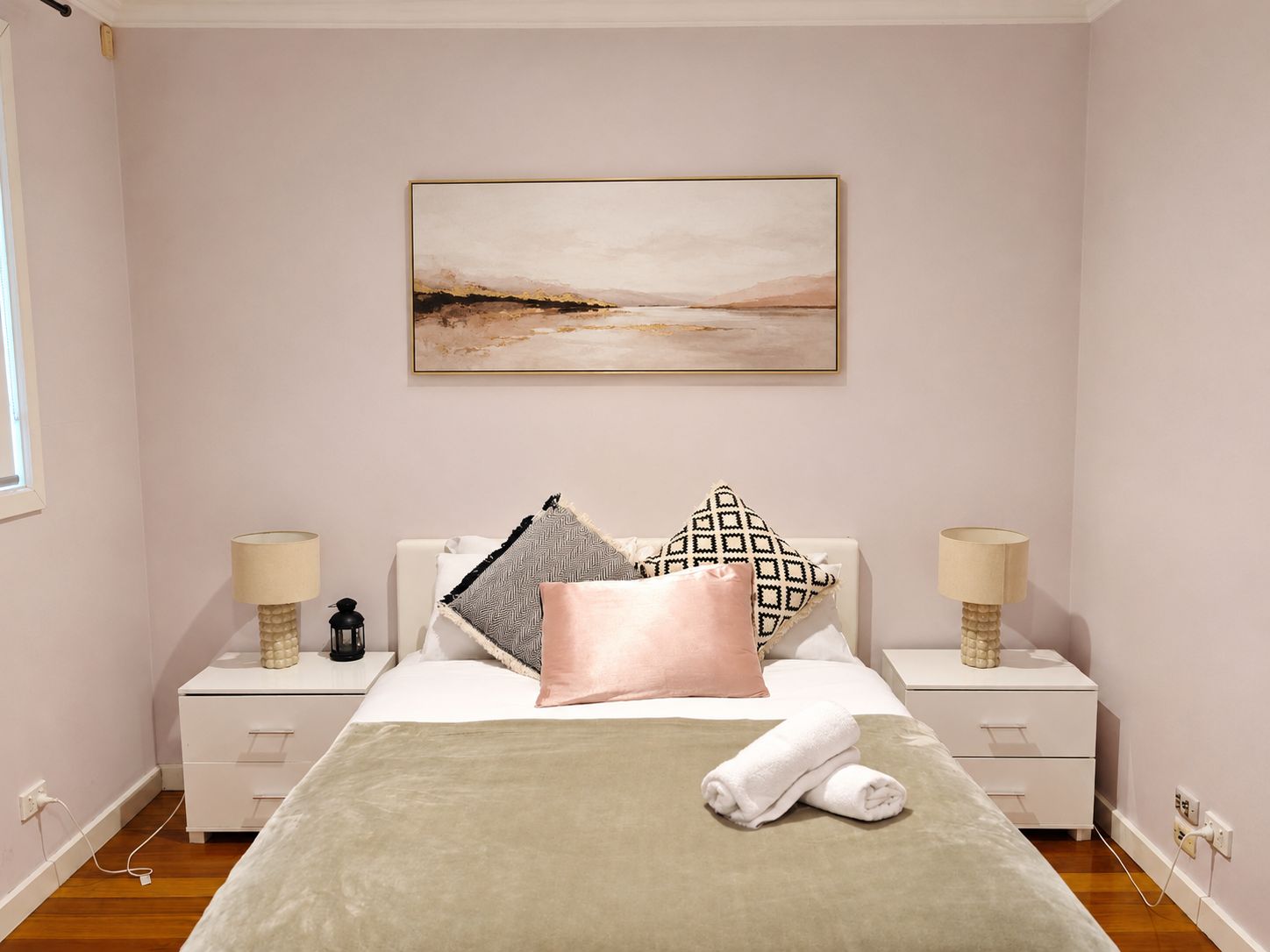 A bedroom with a bed, two bedside tables, a nightstand, a wall-mounted painting, and a window.