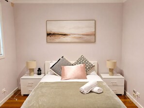 A bedroom with a bed, two bedside tables, a nightstand, a wall-mounted painting, and a window. - Charming 3BR Home with Free Parking in Nunawading (Nunawading)