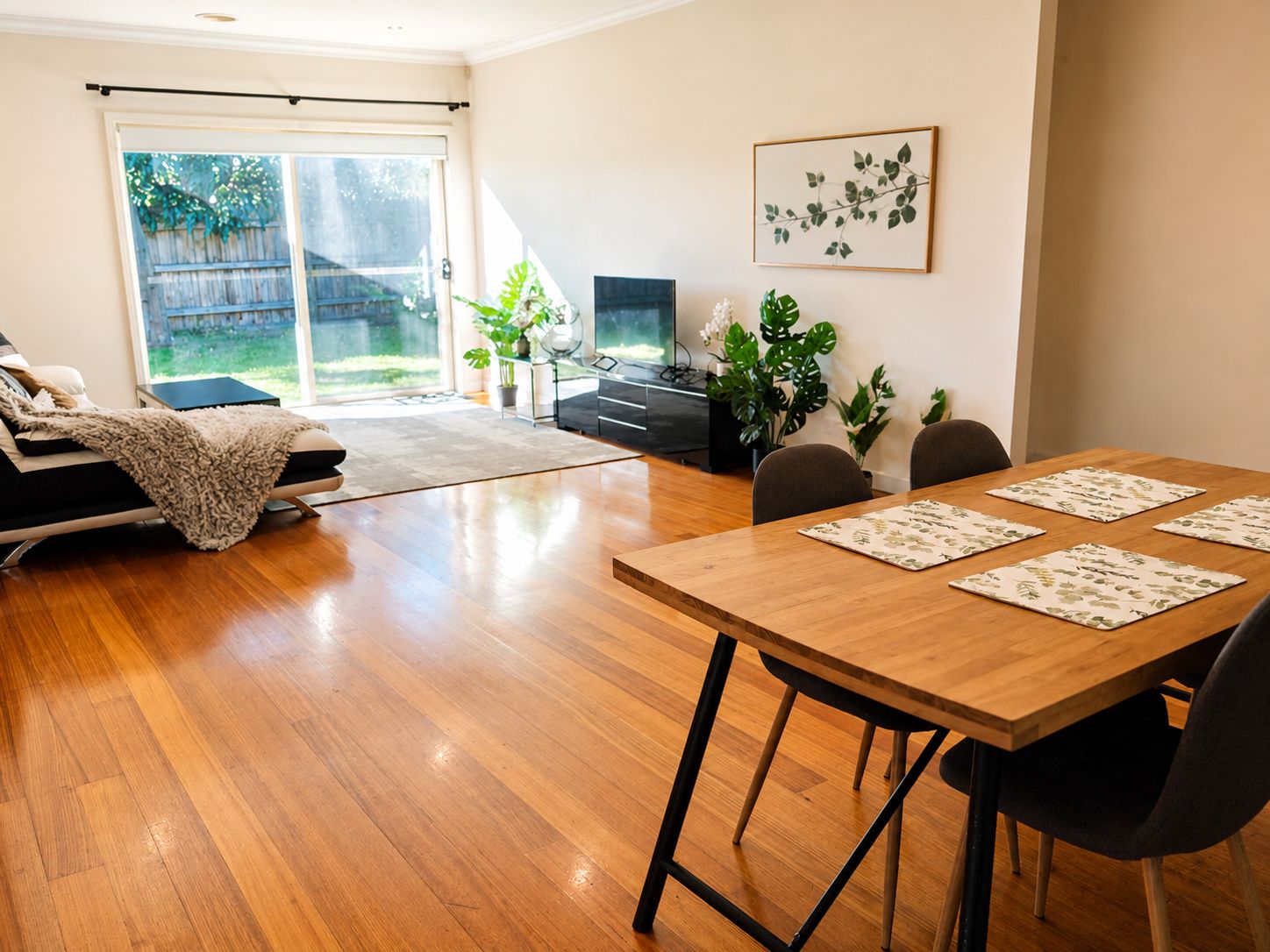 A modern living room with a sofa, a dining table with chairs, a TV, and a large window with a view of a backyard.