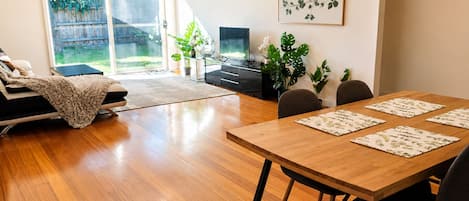 Un salon moderne comprenant un canapé, une table à manger avec des chaises, un téléviseur et une grande fenêtre donnant sur un jardin.