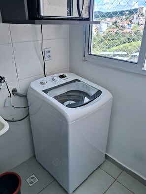 Une petite buanderie équipée d’un lave-linge à chargement frontal, d’une fenêtre donnant sur un quartier résidentiel et d’un sol carrelé.
