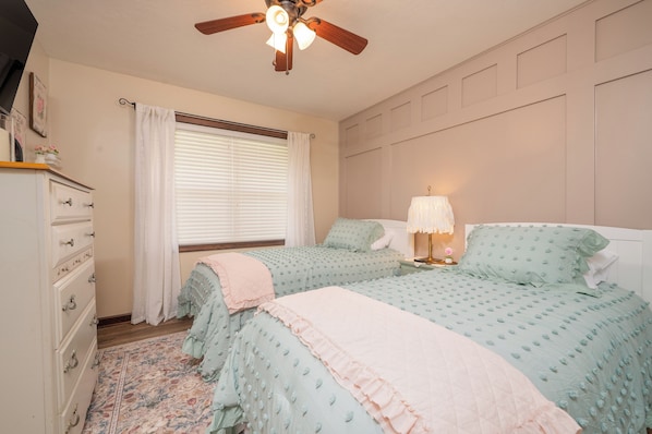 Une chambre avec deux lits, une commode, un ventilateur de plafond et une fenêtre avec des stores.