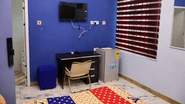 Quarto com parede azul, escrivaninha preta com gavetas, uma cadeira, um frigorífico pequeno e janela com persianas.