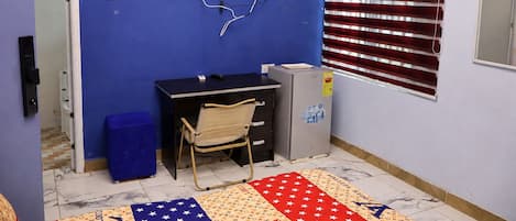 A room with a blue wall, a black desk with drawers, a chair, a small refrigerator, and a window with blinds.
