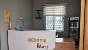 Uma receção com a placa 'Denny's Palace', uma janela com persianas, um frigorífico cheio de bebidas e um aviso de segurança contra incêndios na parede.