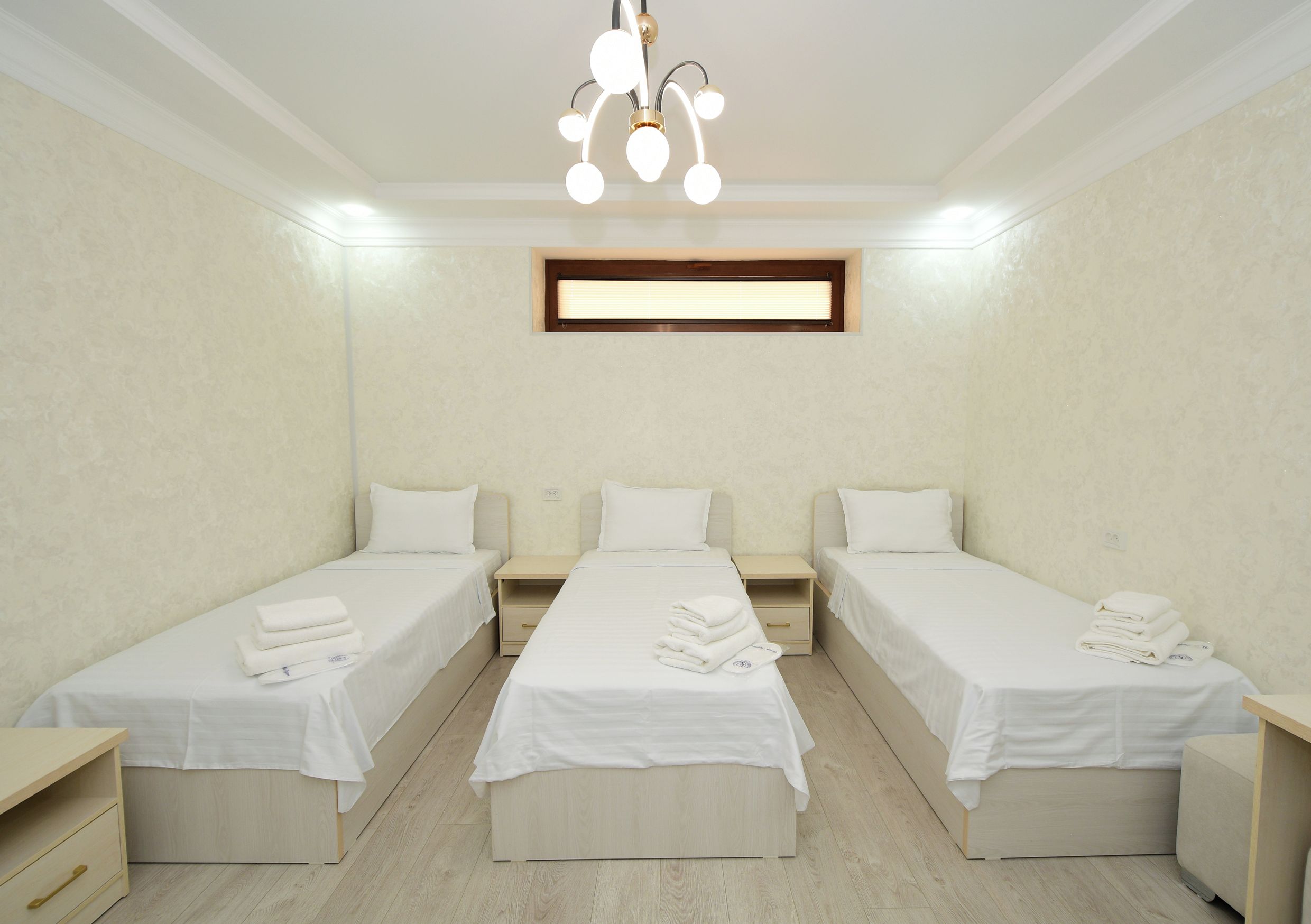 A hotel room with two single beds, a small desk, a nightstand, a ceiling light fixture, and a window with blinds.