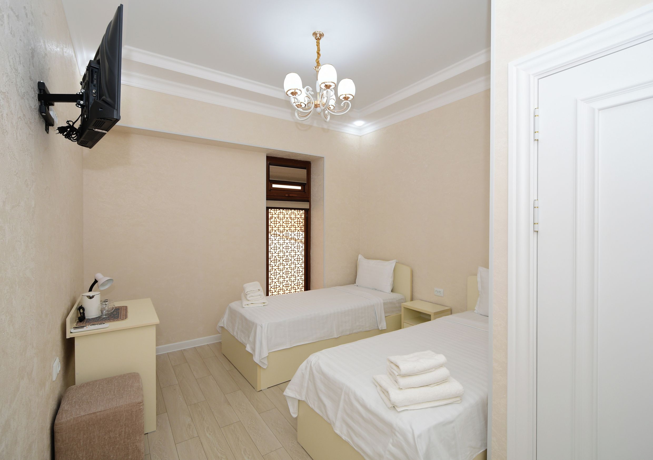 A hotel room with two beds, a desk with a lamp, a TV mounted on the wall, and a chandelier.