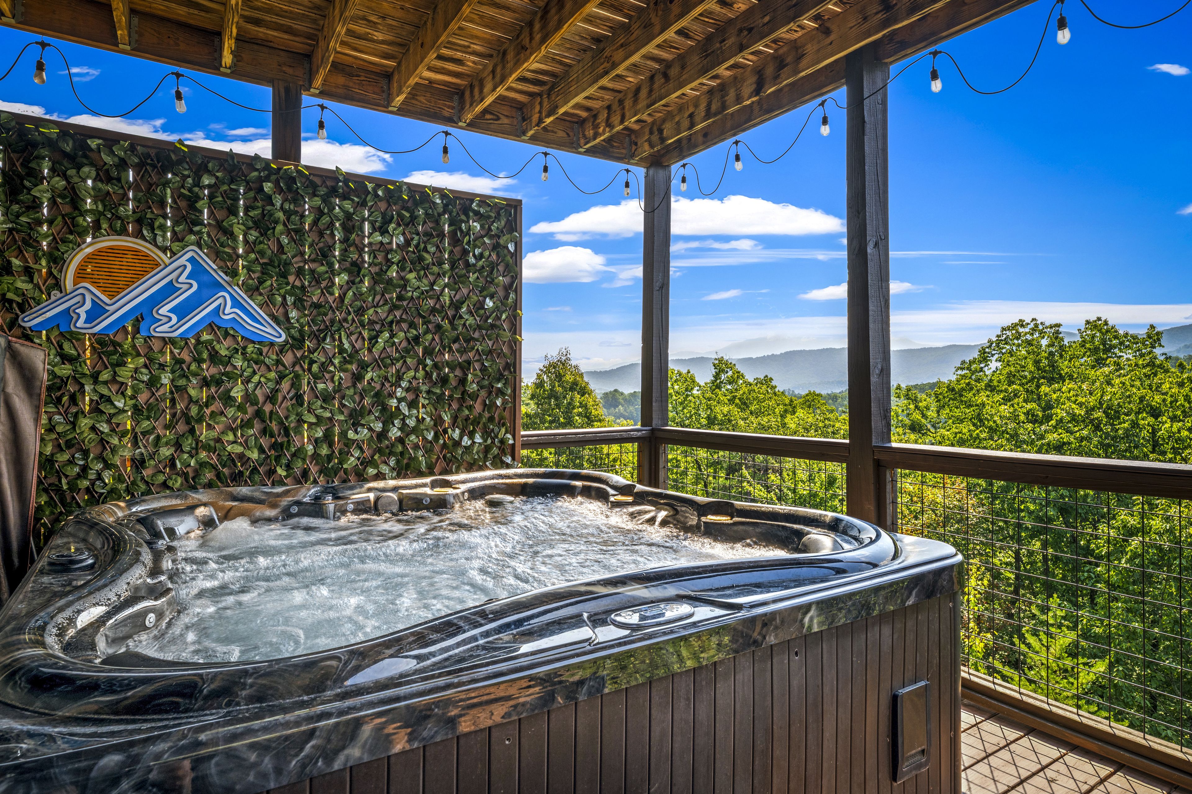 A hot tub with a wooden exterior, surrounded by a wooden deck and a glass railing, overlooking a mountainous landscape.