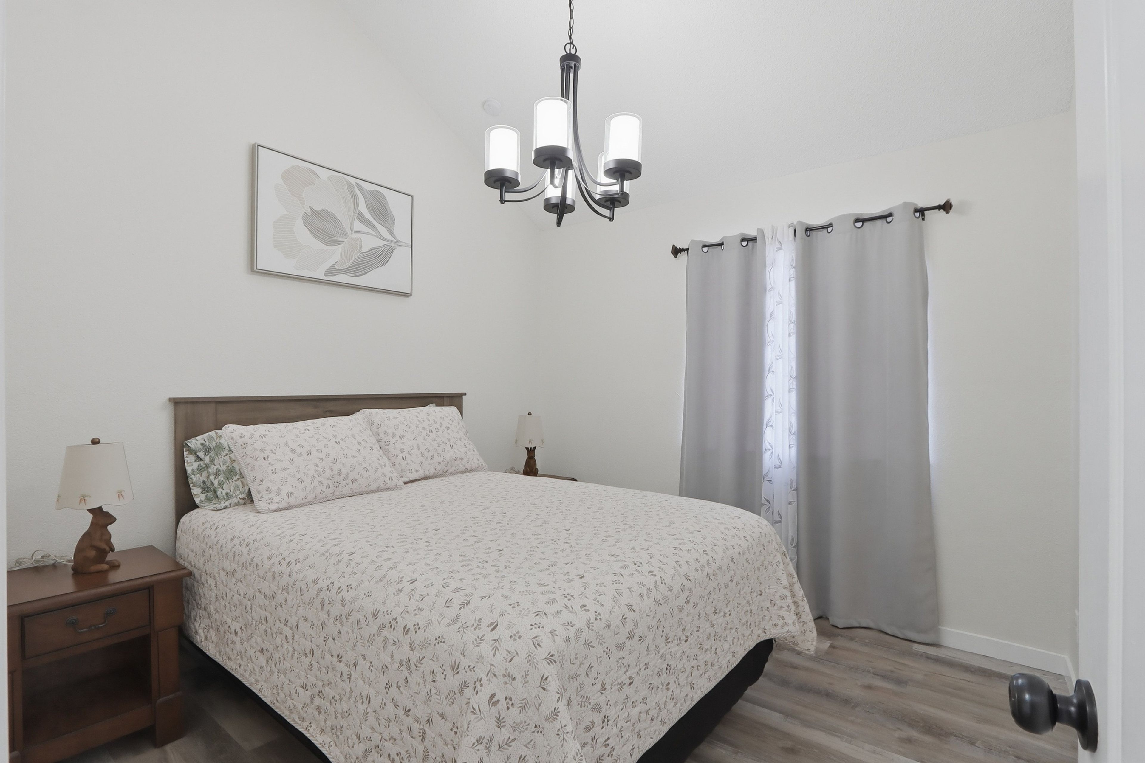 A bedroom with a bed, a nightstand, a lamp, a wall-mounted picture, a chandelier, and a window with curtains.