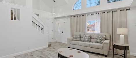 A spacious living area with a high ceiling, a staircase, and a large window.
