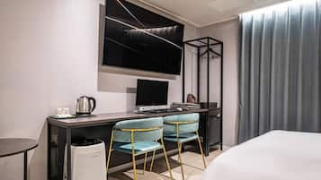 A modern hotel room with a bed, a desk with a computer, two chairs, a TV, and a window with curtains.