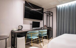 Una habitación de hotel moderna con una cama, un escritorio con computadora, dos sillas, un televisor y un ventanal con cortinas.