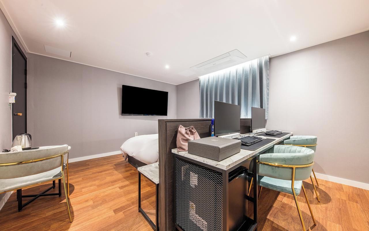A modern hotel room with a bed, a desk with a computer, a TV, and a seating area with chairs.