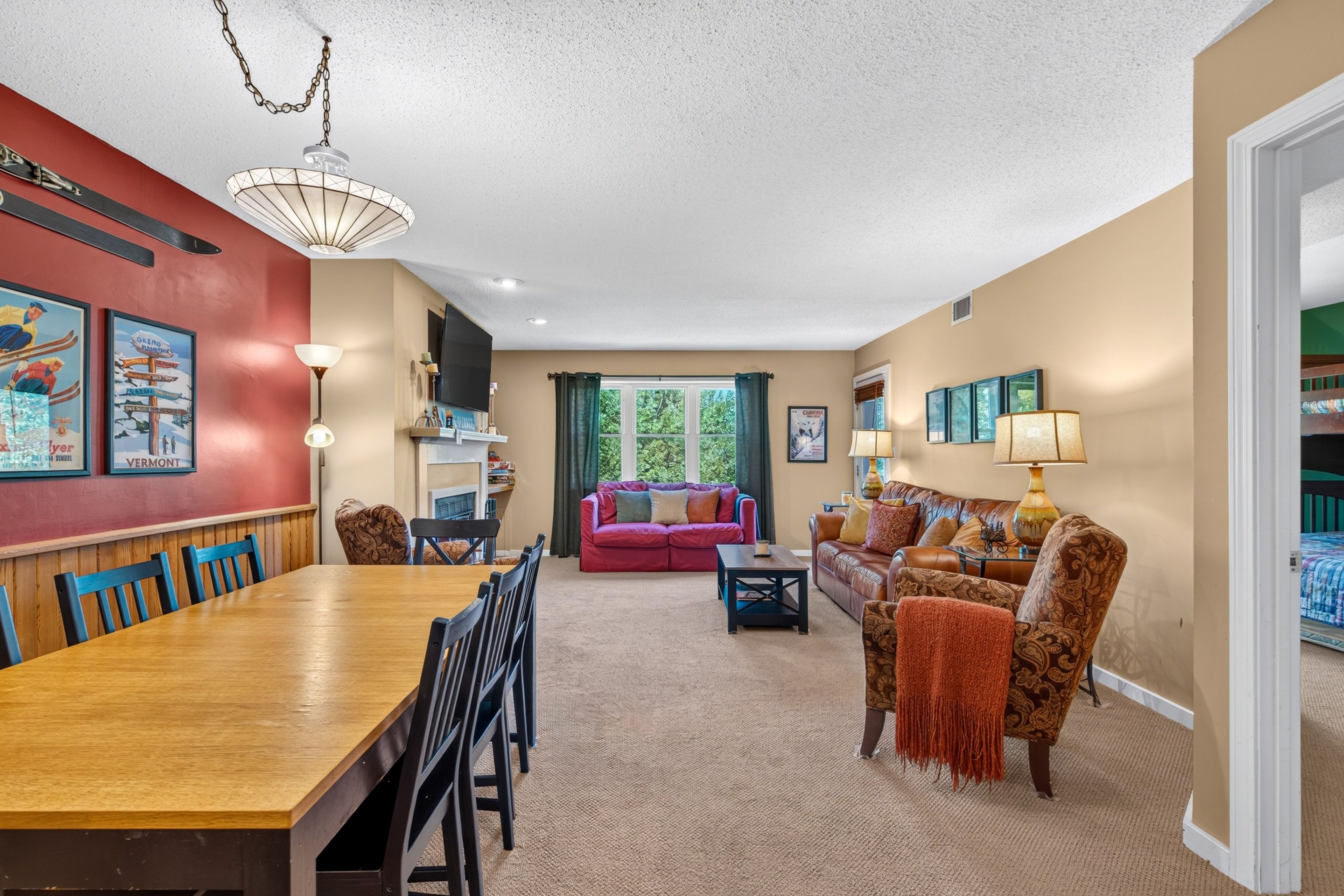 A spacious living area with a dining table, chairs, a sofa, and a coffee table. There is a large window with curtains, a chandelier, and wall art.