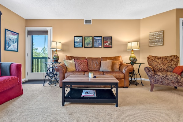 A living room with a brown leather sofa, a red armchair, a patterned armchair, a coffee table, and a small table with a lamp. There are four framed pictures on the wall and a sign with a motivational quote. - White Loop Condo 2 Bedroom Condo (Ludlow)
