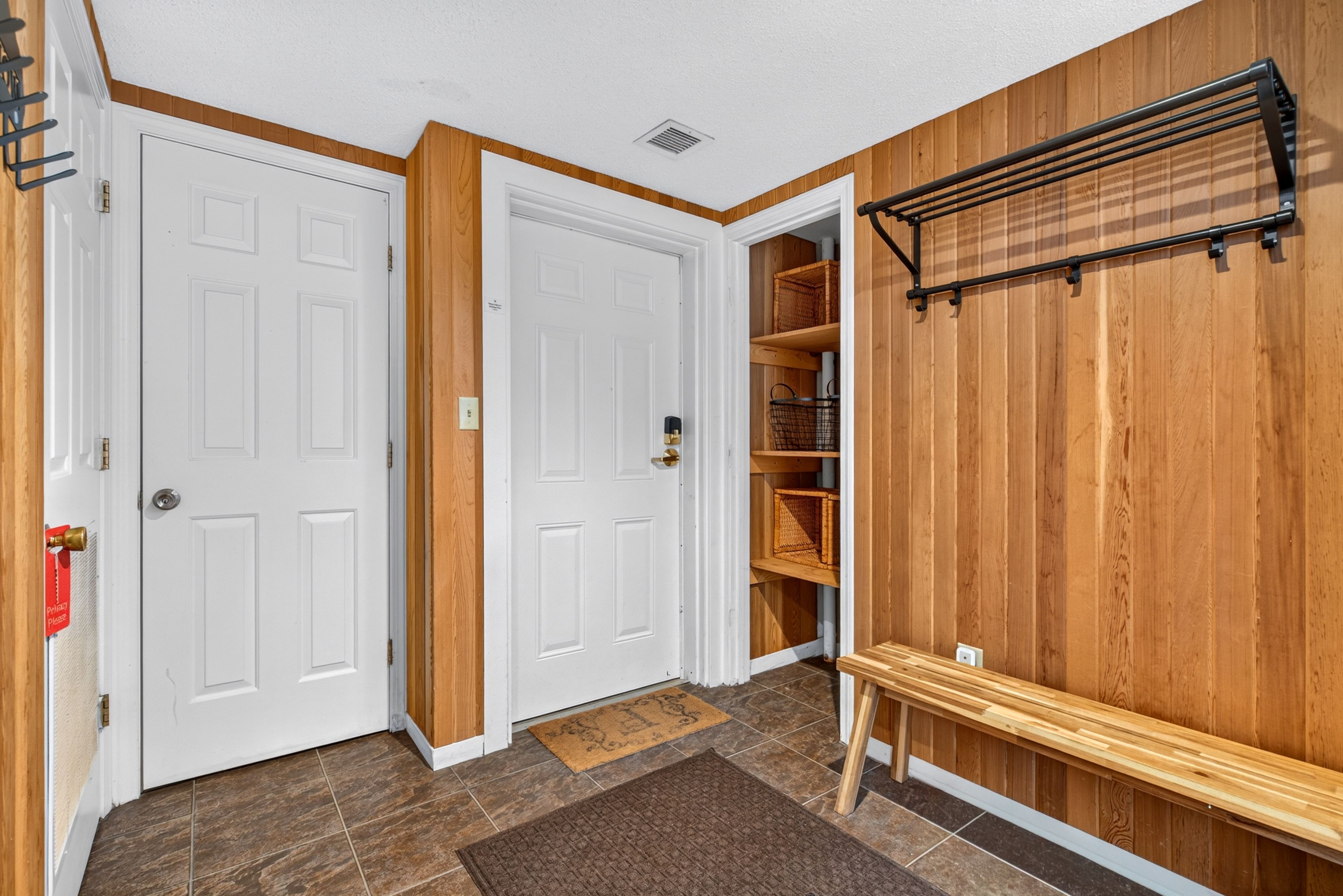 A hallway with two white doors, a wooden bench, a shelf with baskets, and a wall-mounted rack.