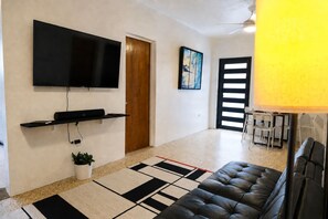 Een woonkamer met een flatscreen televisie, een bank, een bijzettafeltje, een kleine plant en een deur die naar een andere kamer leidt.