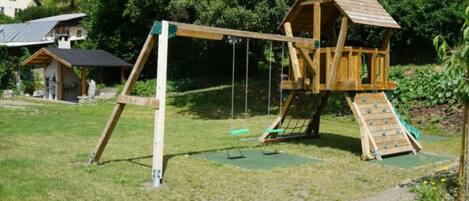 Una estructura de juego de madera con tobogán y columpios, ubicada en un área verde con una casa y árboles al fondo.
