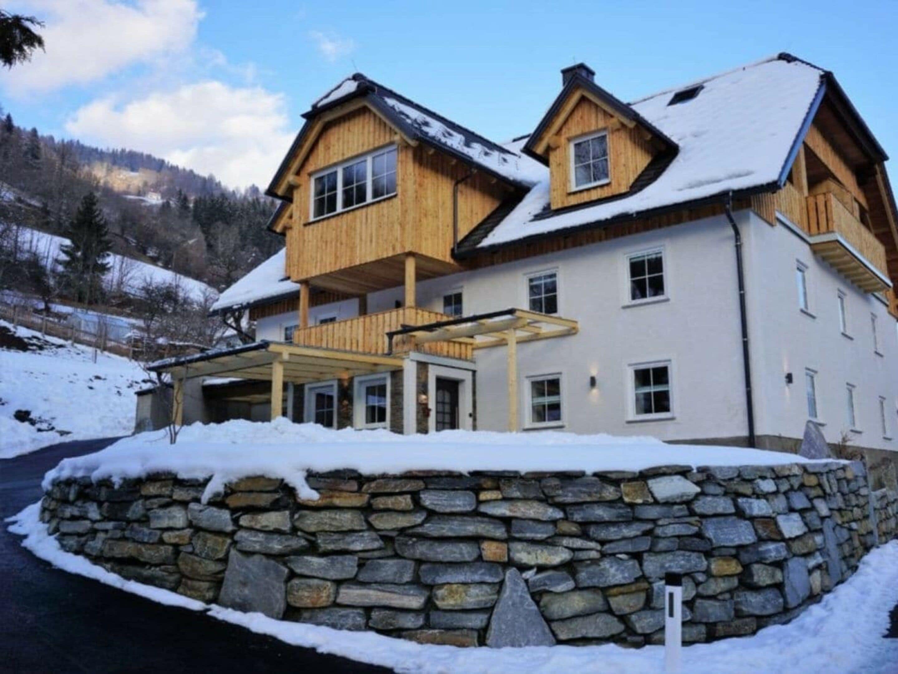 Ein zweistöckiges Holzhaus mit einer Mauer aus Stein davor, umgeben von Schnee.
