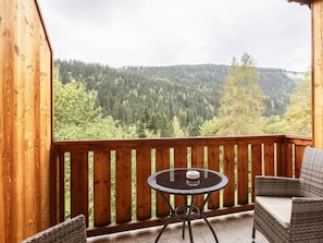 A wooden balcony with a round table and two wicker chairs, overlooking a forested valley.